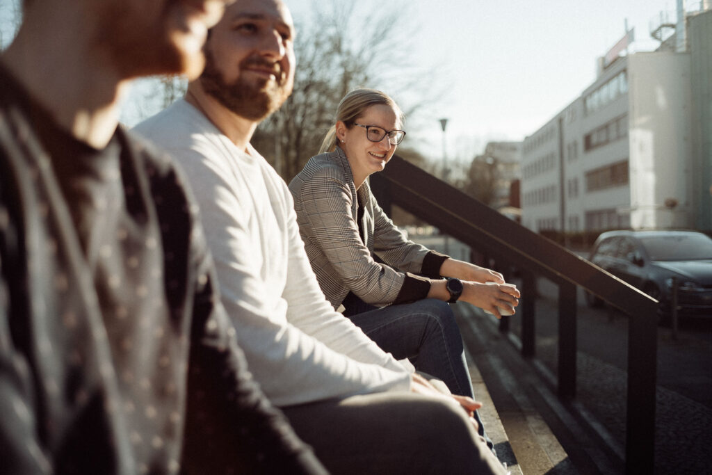 Zwei Männer und eine Frau sitzen draußen vor einem Bürogebäude in der Sonne.
