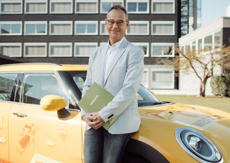 Ein Mann steht vor einem Bürogebäude und lehnt sich an ein WestLotto Auto an.
