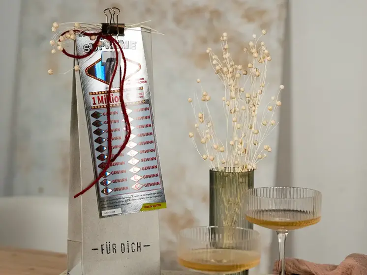 Geschenkverpackung mit angeklammertem Rubbellos Supreme 7, daneben zwei Cocktails und eine Vase mit Trockenblumen.