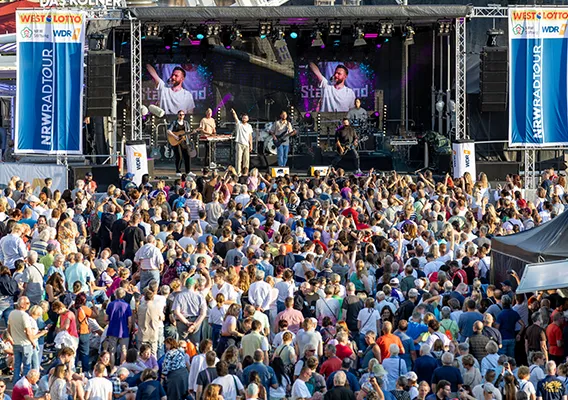Menschen feiern vor einer Buehne auf der eine Band spielt. Abendveranstaltung der NRW-Radtour.