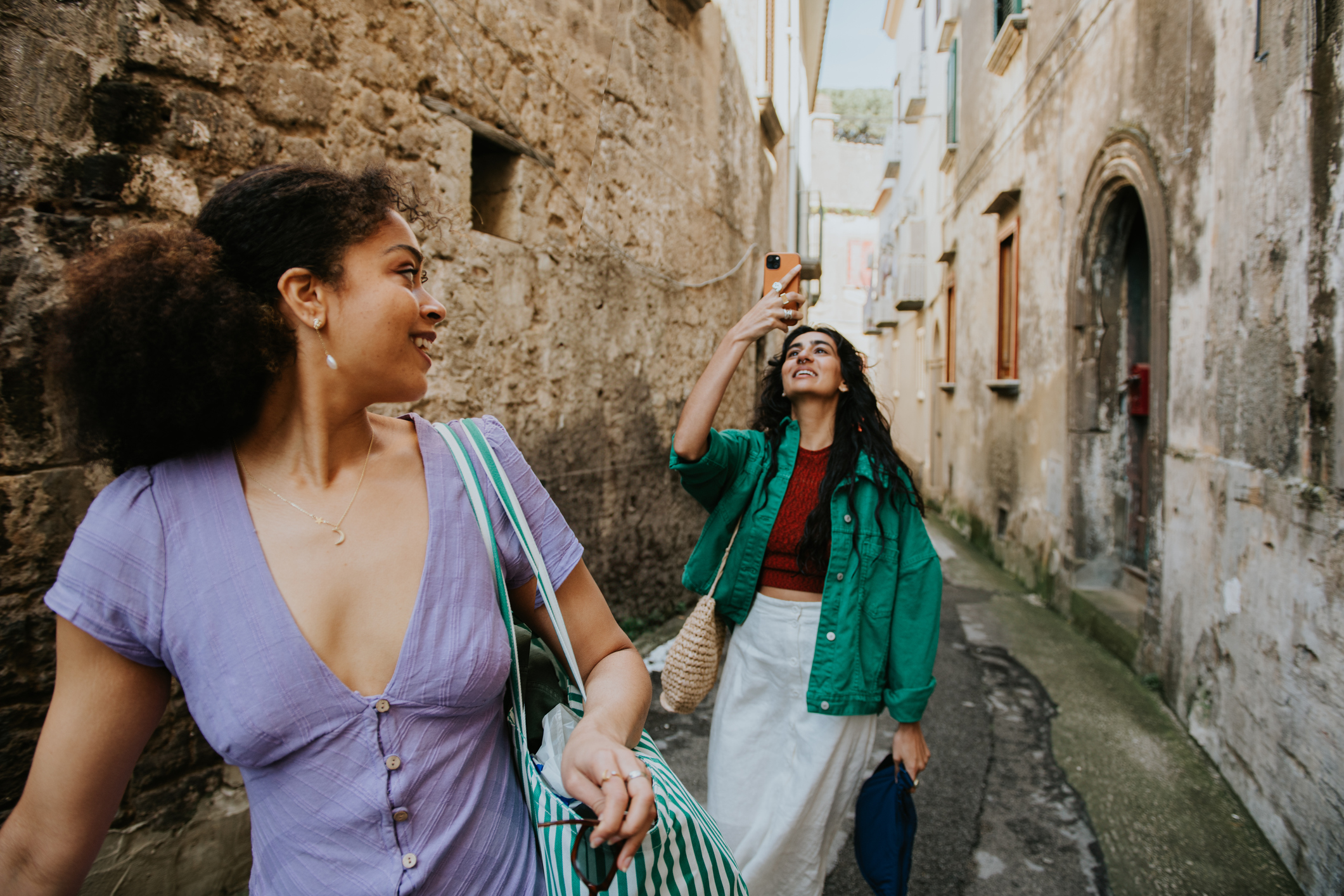 Zwei Frauen sind in einer hellen Gasse umgeben von alten Gemäuern unterwegs. 