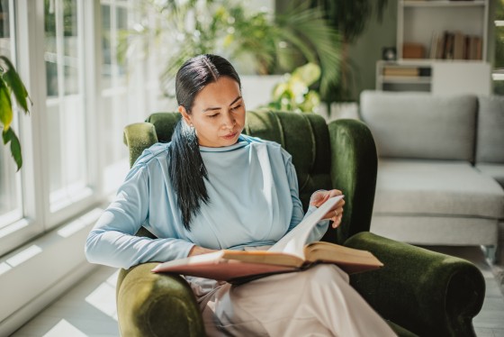 Eine Frau liest ein Buch auf einem grünen sessel in einem hellen Wohnzimmer.