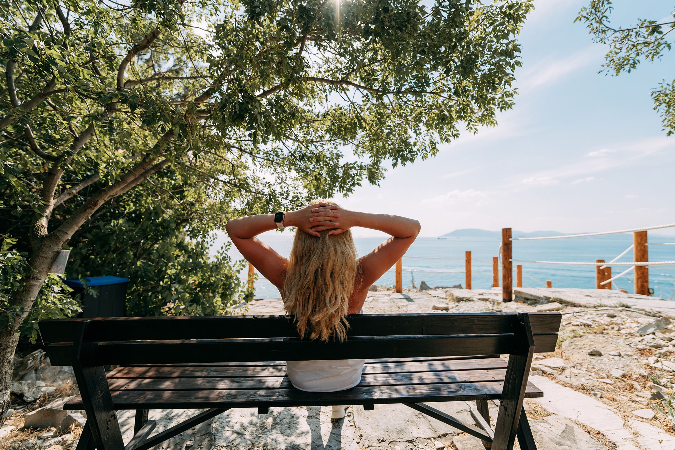 Junge Frau auf einer Bank am Meer, mit dem Ruecken zum Betrachter.