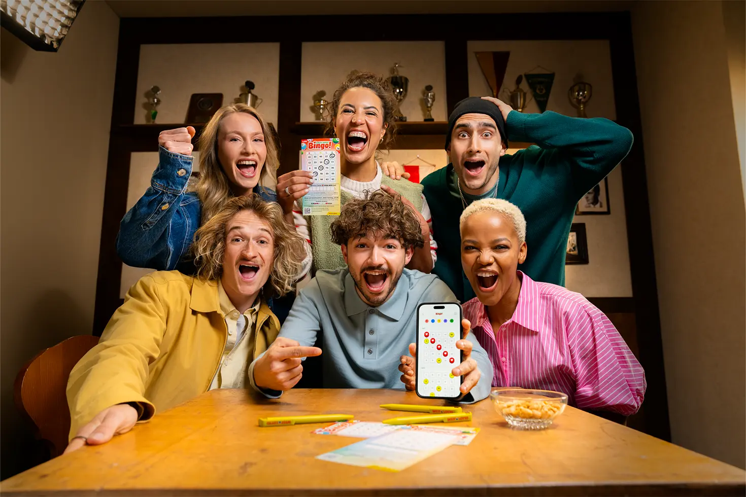 Mehrere Personen spielen gemeinsam Bingo! und zeigen begeistert ein Bingo!-Los sowie das Bingo!-Los auf einem Smartphone.
