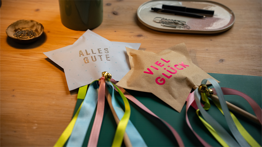 Zwei gebastelte Sterne mit Satinbändern und kleinen Glöckchen an einem Holzstock - WestLotto DIY-Idee