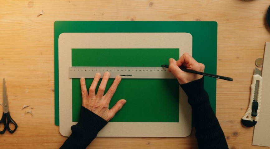 Rubbellos-DIY: Zwei Hände zeichnen mit Hilfe eines Lineals auf einem grünen Pappkarton