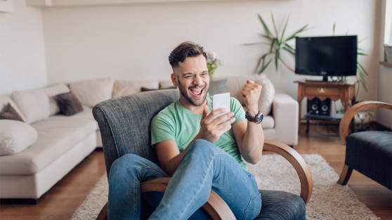 Mann sitzt auf Sessel, schaut auf Handy und freut sich | Gewinner WestLotto-Rubbellos