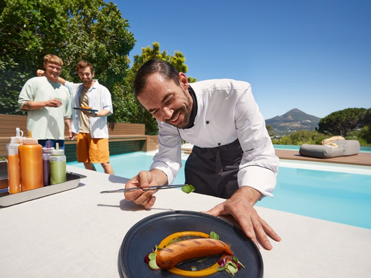 Ein Koch steht vor einem Pool und verziert eine Bratwurst | Lotterie Eurojackpot