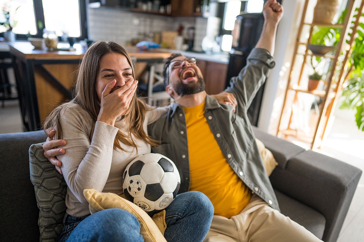 Eine Frau und ein Mann sitzen mit einem Fußball auf dem Sofa und jubeln - WestLotto TOTO Sondersauslosung