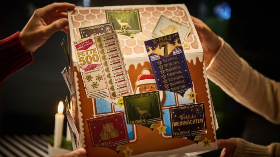 Vier Hände halten einen Rubbellos-Adventskalender in Lebkuchenform über einem Tisch - WestLotto