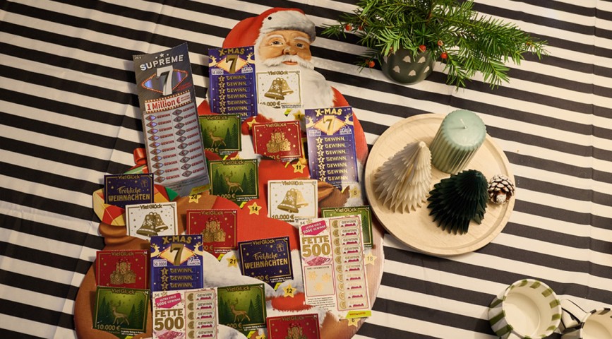 Ein Rubbellos-Adventskalender in Weihnachtsmann-Form liegt auf einem weihnachtlich dekoriertem Esstisch - WestLotto