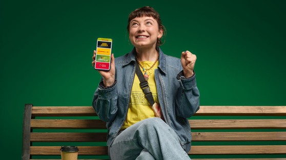 Frau sitzt auf einer Parkbank und hat ein Smartphone in der Hand, auf dem die WestLotto-App geöffnet ist. | WestLotto