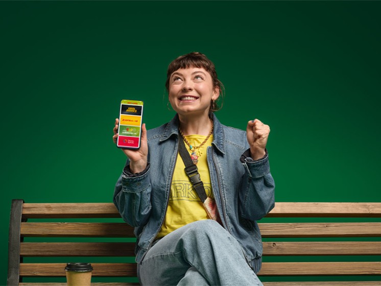 Frau sitzt auf Parkbank, hält Smartphone mit geöffneter WestLotto-App in der Hand und freut sich | WestLotto