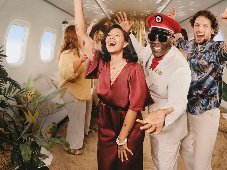 Drei Personen tanzen in einem Flugzeug, dessen Boden mit Sand bedeckt ist. Hinter ihnen weitere tanzende Menschen - Westlotto