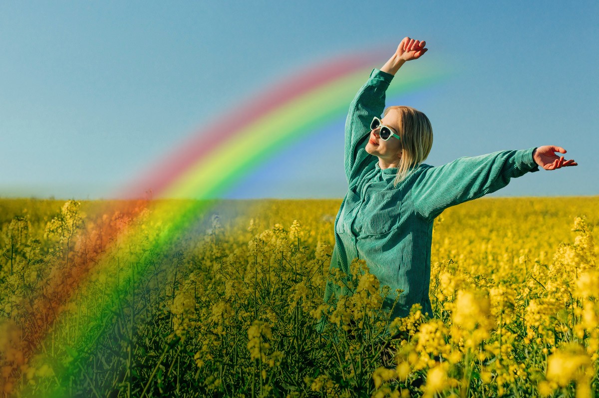 Frau steht in Blumenfeld und streckt Hände in die Luft. Davor ein Regenbogen - WestLotto GlücksSpirale