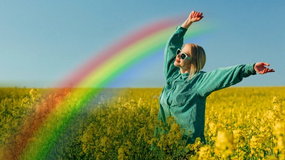 Frau steht in Blumenfeld und streckt Hände in die Luft. Davor ein Regenbogen - WestLotto GlücksSpirale