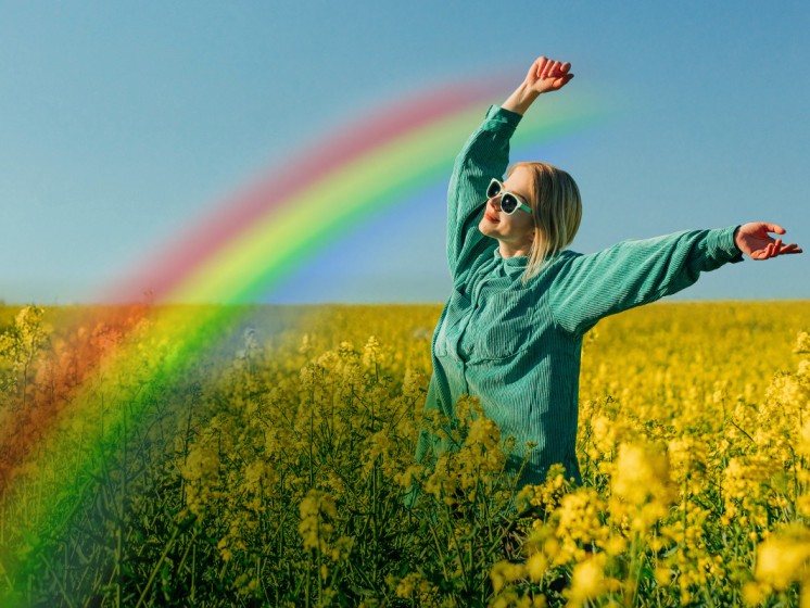 Frau steht in Blumenfeld und streckt Hände in die Luft. Davor ein Regenbogen - WestLotto GlücksSpirale