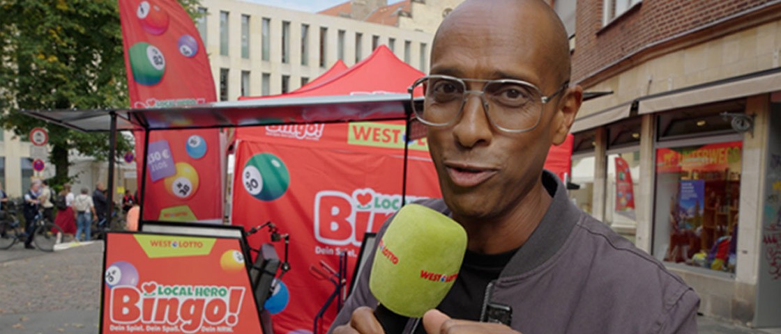Moderator Amiaz Habtu mit Mikrofon in der Hand steht vor einer Rikscha - WestLotto Local Hero Bingo