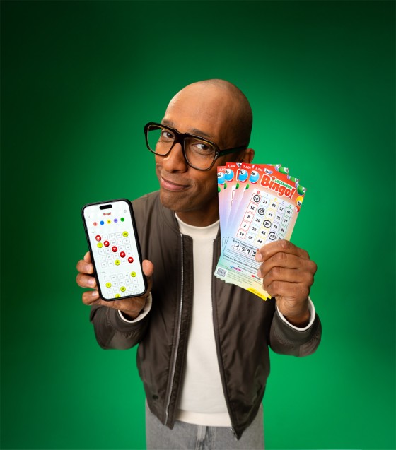 Amiaz Habtu - Moderator der Local Hero Bingo!-Show - hält Bingo!-Lose und Smartphone hoch. WestLotto