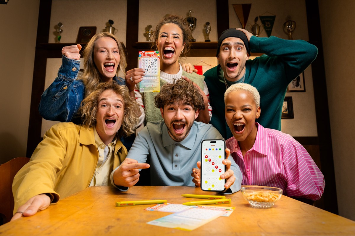 Gruppe von Menschen in einer Kneipe spielen Local Hero Bingo! mit App und Spielschein. WestLotto