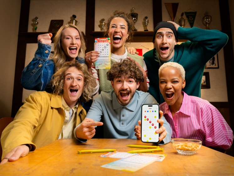 Gruppe von Menschen in einer Kneipe spielen Local Hero Bingo! mit App und Spielschein. WestLotto