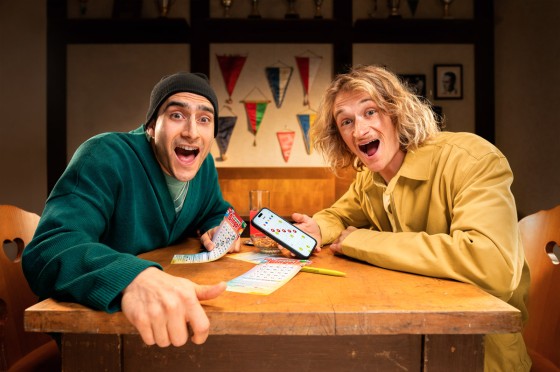 Zwei Männer in einer Kneipe spielen Local Hero Bingo! mit App und Spielschein. WestLotto