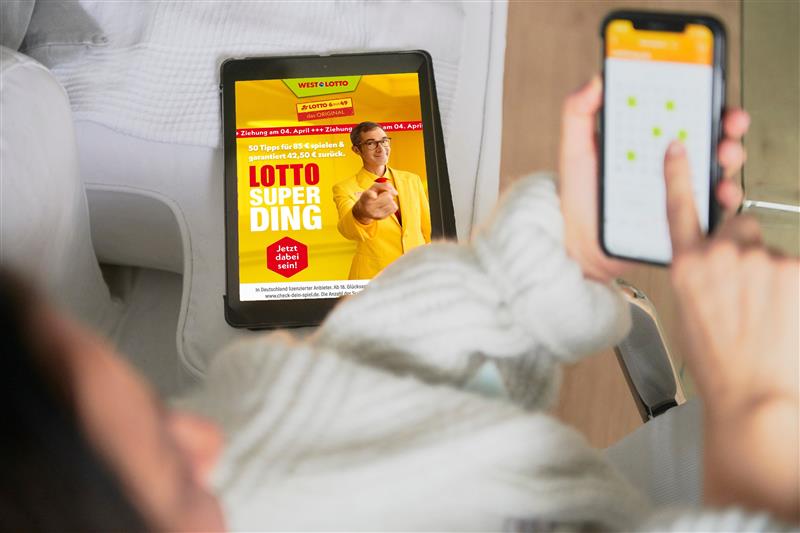 Person sitzt auf Couch und hält Handy in der Hand. Daneben liegt ein Tablet, auf welchem eine Anzeige für das Lotto-SuperDing abgebildet ist. 