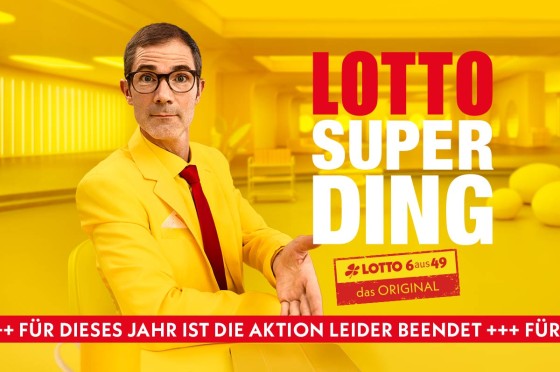 Mann mit gelbem Anzug steht vor gelbem Hintergrund und zeigt mit Hand auf Uhr - WestLotto Lotto-SuperDing