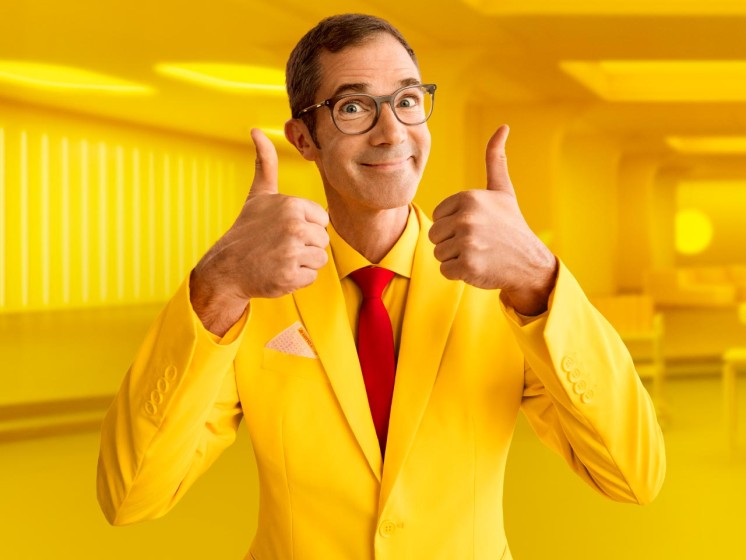 Mann mit gelbem Anzug steht vor gelbem Hintergrund und hält beide Daumen nach oben - WestLotto Lotto-SuperDing