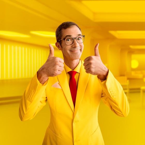 Mann mit gelbem Anzug steht vor gelbem Hintergrund und hält beide Daumen nach oben - WestLotto Lotto-SuperDing