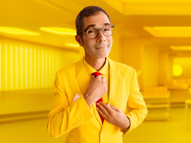 Mann mit gelbem Anzug steht vor gelbem Hintergrund und richtet seine Krawatte - WestLotto Lotto-SuperDing