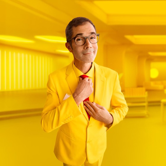 Mann mit gelbem Anzug steht vor gelbem Hintergrund und richtet seine Krawatte - WestLotto Lotto-SuperDing