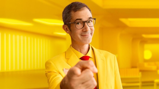 Mann mit gelbem Anzug steht vor gelbem Hintergrund und zeigt mit Finger auf Betrachter - WestLotto Lotto-SuperDing