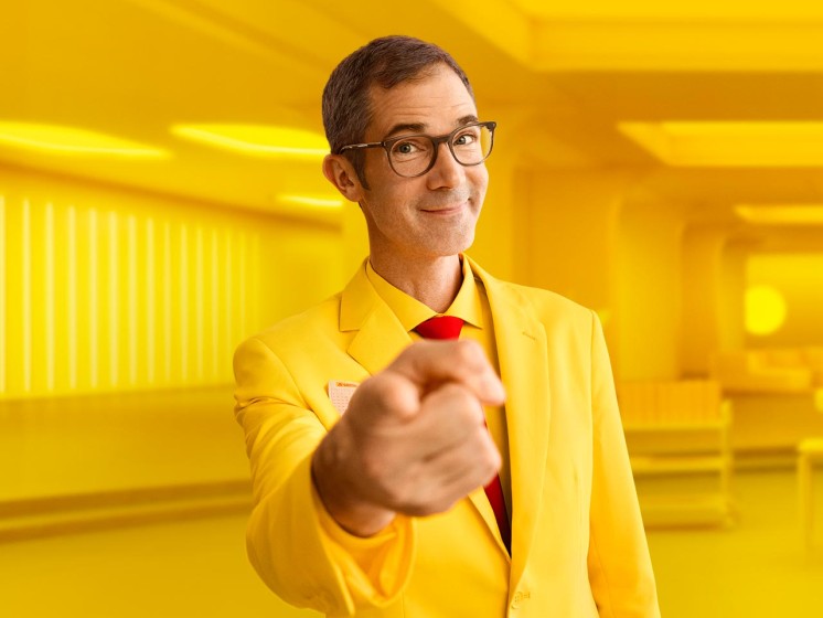 Mann mit gelbem Anzug steht vor gelbem Hintergrund und zeigt mit Finger auf Betrachter - WestLotto Lotto-SuperDing