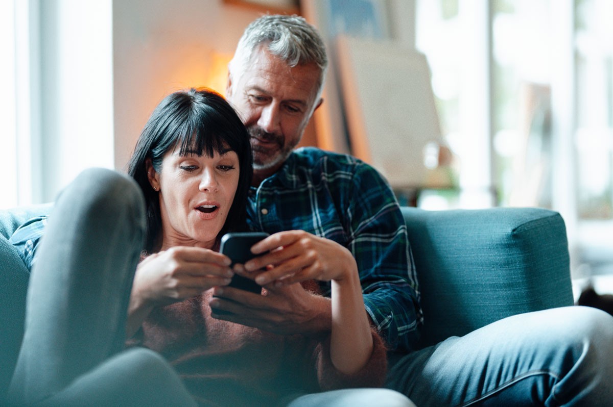 Pärchen sitzt auf Couch und schaut erstaunt auf Smartphone - WestLotto