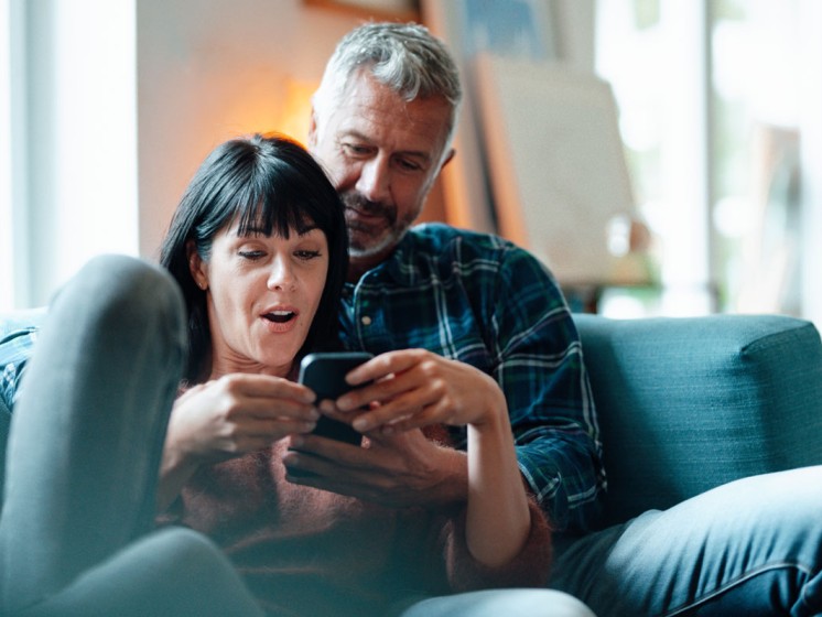 Pärchen sitzt auf Couch und schaut erstaunt auf Smartphone - WestLotto