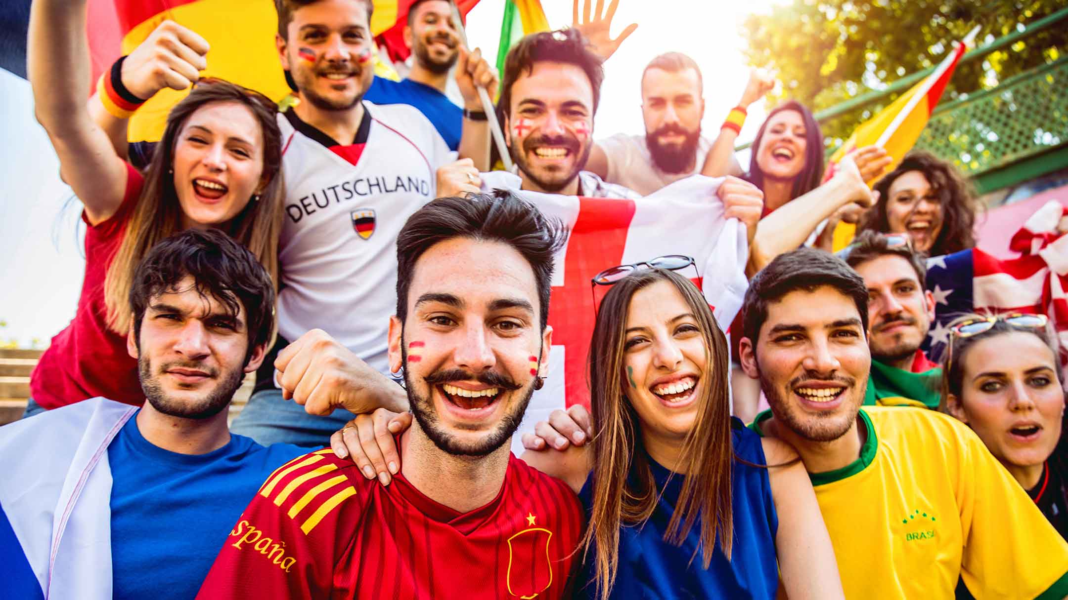 Gruppe von Fußballfans in Fantrikots verschiedener Länder jubelt gemeinsam und hält Fahnen hoch.
