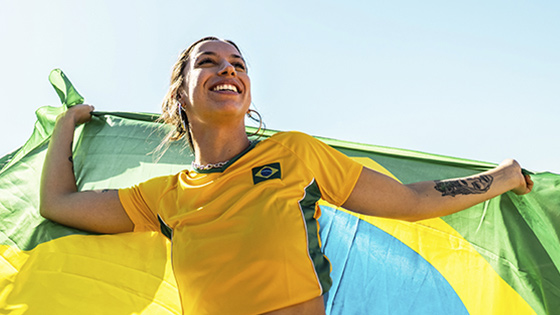 Person mit ausgebreiteten Armen vor einer brasilianischen Flagge im Hintergrund, gekleidet in ein gelbes Trikot, im Freien bei klarem Himmel.